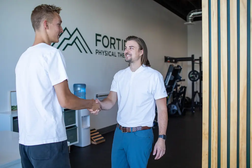 A Fortify physical therapist shaking hands with a patient in the lobby, under the painted Fortify Physical Therapy wall mark
